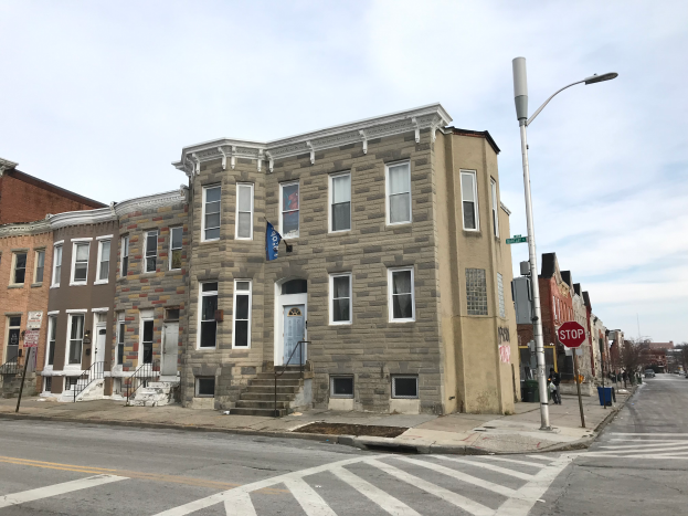Eine Reihe neuer Häuser an einer Straßenecke in Philadelphia, mit sichtbaren Fenstern, Türen, Treppen mit Geländern, Laternen, Schildern und Fahrzeugen auf der Straße, mit Bäumen und einem bewölkten Himmel im Hintergrund.