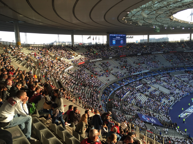 Eine große Menschenmenge sitzt im Allianz Arena Stadion in München, Deutschland, und schaut ein Fußballspiel, mit einer Bühne auf der rechten Seite, Fahnen, Stangen und einem Bildschirm im Hintergrund und dem Himmel oben sichtbar.