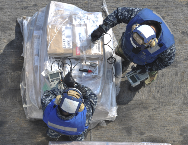 Zwei Männer in blauen Uniformen und Helmen arbeiten an einem großen Drohnen, hältend verschiedene Gegenstände, während eine Plastikabdeckung auf dem Boden vor ihnen liegt.