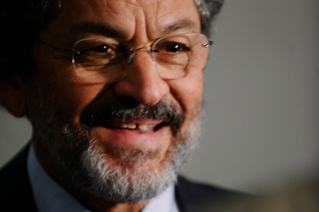 Ein älterer Mann in einem schwarzen Anzug und weißem Hemd, der eine Brille trägt, lächelt.