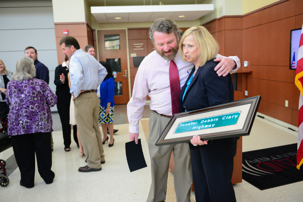 Eine Gruppe von Menschen, die drinnen stehen, einer hält einen Foto Rahmen mit 'Senator Debbie Clary Highway' darauf, mit einer Flagge, einer Wand, einer Tür und Lichtern im Hintergrund.