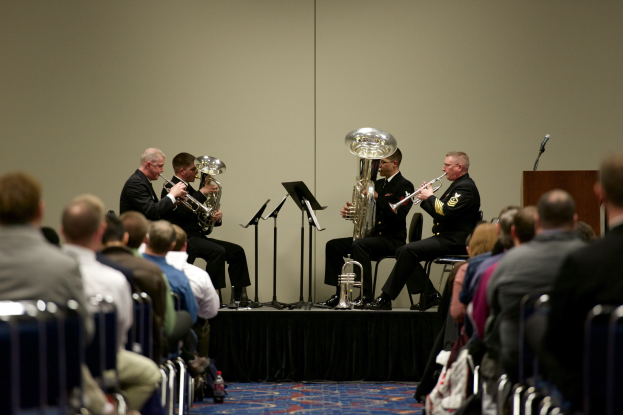 Gruppe von Männern, die auf einer Bühne Blasinstrumente spielen, mit Notenständern, einem Podium mit Mikrofon und einem Publikum, das auf Stühlen sitzt, mit einer Wand im Hintergrund.