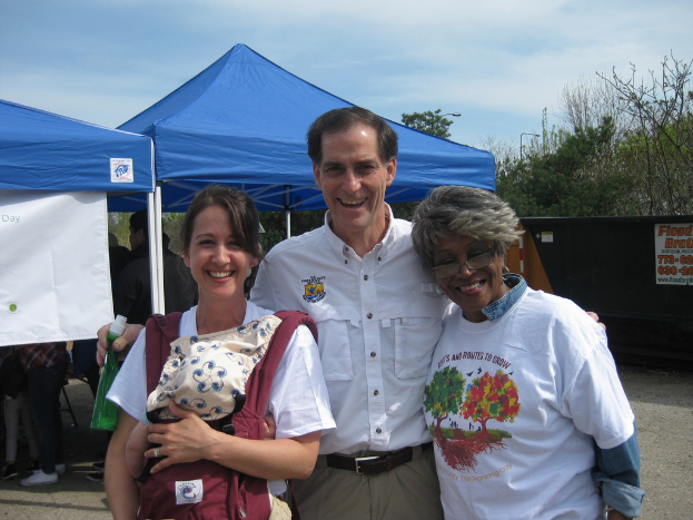 Eine Gruppe von Menschen, die unter Zelten steht und lacht, mit einer Frau, die ein Baby hält, einem Mann, der eine Flasche hält, Bäumen und einem blauen bewölkten Himmel im Hintergrund.