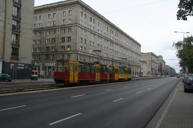 Eine Stadtansicht mit Gebäuden, Fahrzeugen, einem Zug, Straßenlaternen, Pfählen, Kabeln, einer Straße, Bäumen, Fenstern, Geschäften und einer bewölkten Himmel.