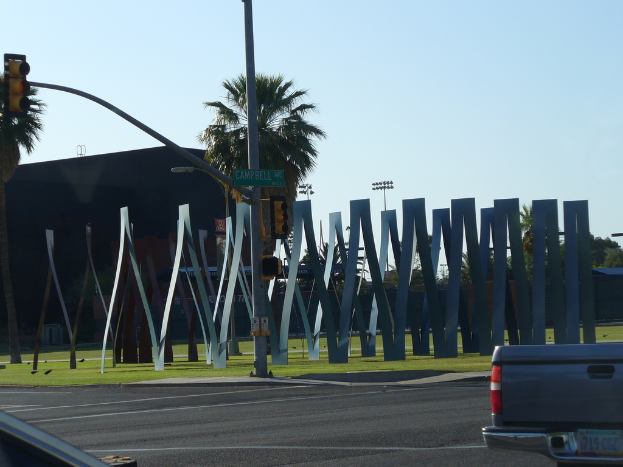 Eine Straßenszene mit Straßenlaternen, Ampelmasten, einem Schild, Metallstangen, Gras, Gebäuden, Bäumen und einem sichtbaren Kofferraum in der unteren rechten Ecke unter dem Himmel.