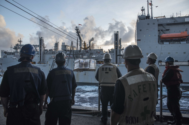 Gruppe von Männern in Helmen, die auf einem Boot mit Relings stehen, umgeben von Schiffen, Pfählen und Drähten im Hintergrund unter einem bewölkten Himmel.