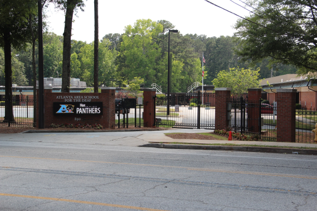 Einfahrt zu einer Schule mit einem Schild, umgeben von Bäumen, Straßenmöbeln, Fahrzeugen und Gebäuden unter einem klaren blauen Himmel.
