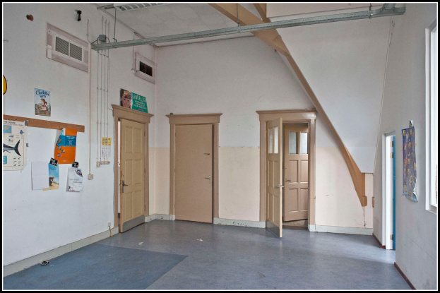 Ein leerer Raum mit vielen Fenstern und Türen, ein Anschlagbrett mit Papieren an der linken Wand, Plakate an der rechten Wand, Rohre und eine Klimaanlage an der Decke und Text am unteren Bildrand.