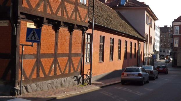Fahrzeuge auf der Straße mit Gebäuden, Laternenmasten, Werbetafelmasten, Fenstern und Dächern im Hintergrund und einem Fahrrad auf dem Gehweg in der Nähe eines Mastes.