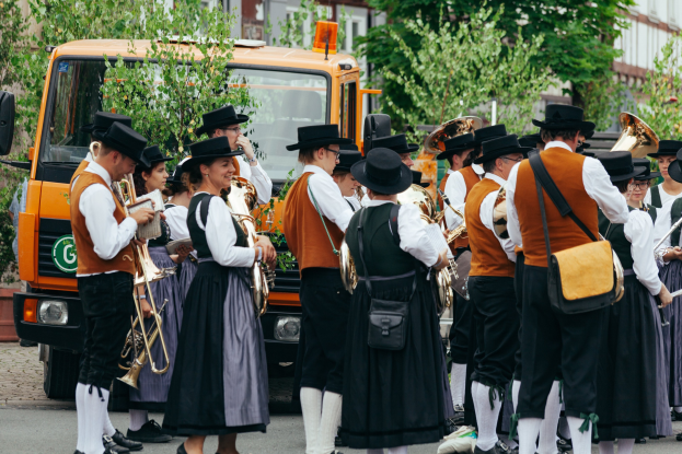 Eine Gruppe von Menschen in traditioneller bayrischer Tracht, die eine Straße entlangmarschieren, Instrumente spielen und Taschen tragen, mit Bäumen und Gebäuden im Hintergrund und einem geparkten Fahrzeug.