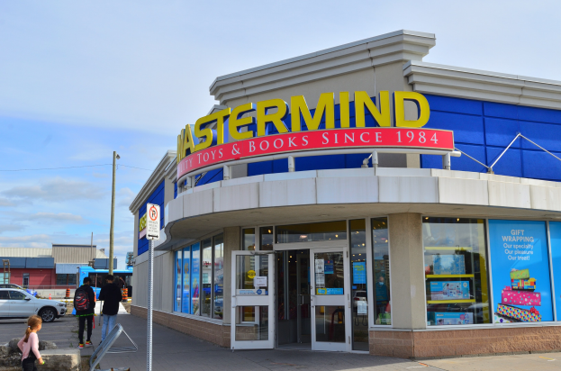 Vorderseite von "Ostermind-Spielwaren & Bücher" mit einer Gruppe von Menschen davor, Fahrzeugen auf der Straße, einem Wegweiser und einem bewölkten Himmel im Hintergrund.