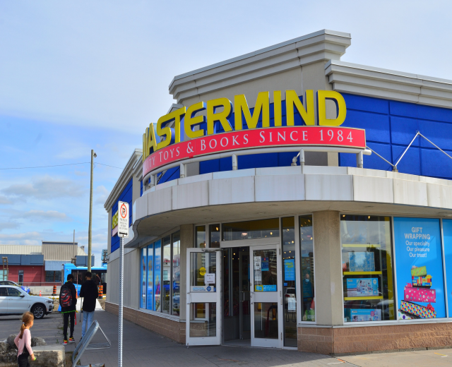 Vorderseite von "Ostermind-Spielwaren & Bücher" mit einer Gruppe von Menschen davor, Fahrzeugen auf der Straße, einem Wegweiser und einem bewölkten Himmel im Hintergrund.