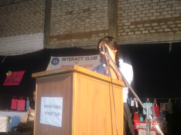 Ein Mädchen in einem weißen Hemd steht näher an einem Rednerpult in einem Konferenzraum, mit einem Mikrofonständer rechts daneben, einem weißen Banner auf einem Hintergrund dahinter und mehreren Taschen, die an einem Faden rechts hängen.