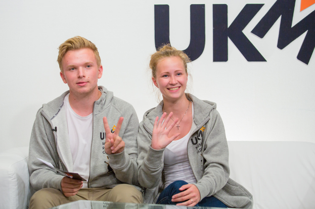 Ein Paar in passenden Jacken sitzt auf einem Sofa mit 'UKM' im Hintergrund.