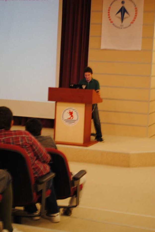 Eine Person steht auf einem Podium, spricht, mit mehreren sitzenden Personen links, einer Tafel und einem Vorhang links und einer weiteren Tafel im Hintergrund.