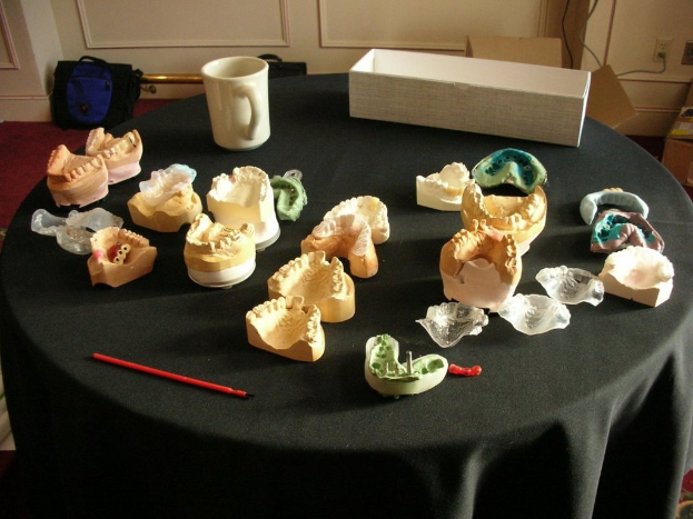 Ein schwarzer Tisch mit einem Becher, einem weißen Kasten und verschiedenen Gegenständen vor einer Wand mit Taschen, Kartons und Stromanschlüssen auf einem Teppichboden.