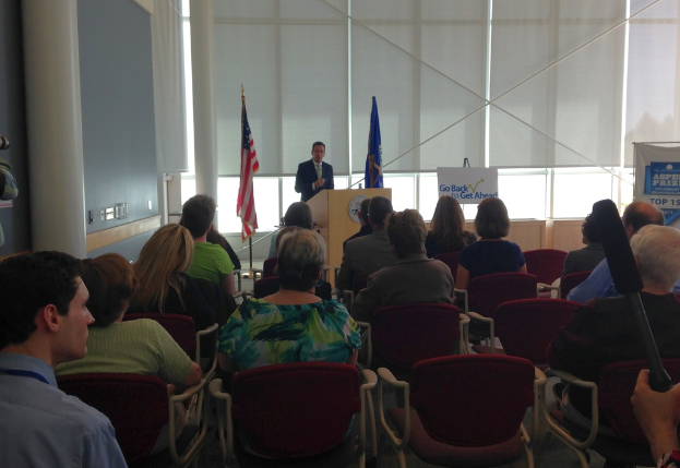 Ein Mann steht an einem Rednerpult und spricht zu einem Publikum, das auf St├╝hlen sitzend zuh├Ârt, mit zwei Fahnen neben ihm und einer Wand, Säulen und Fenstern im Hintergrund.