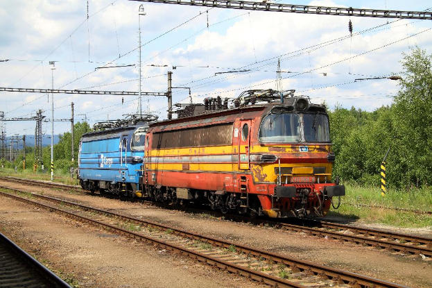 Eine Lokomotive auf Schienen mit Strommasten, Kabeln, Bäumen, Pflanzen und Wolken am Himmel.