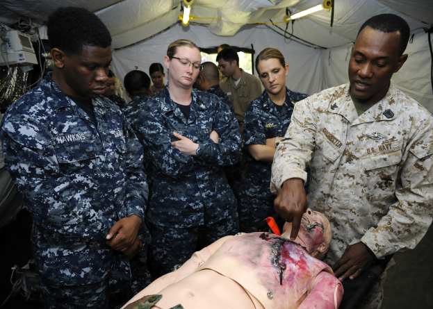 Eine Gruppe von Menschen in Militäruniformen umgibt einen Mann auf einer Trage, wobei eine Person ein Objekt hält und ein beleuchtetes Zelt im Hintergrund zu sehen ist.