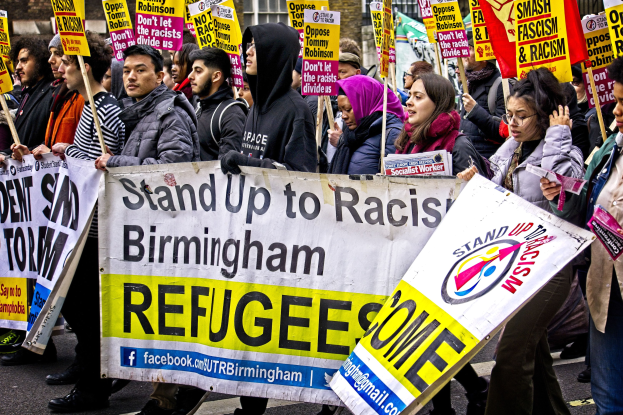 A diverse group of people marching down a street, holding protest signs and banners in front of a building with windows.