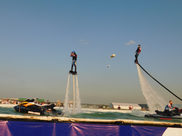 Group of people riding jet skis on water with a banner below, against a backdrop of buildings, people, and a clear blue sky.