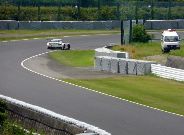 Ein Rennwagen fährt auf einer Rennstrecke mit einem Polizeiwagen im Hintergrund, umgeben von Grün und Absperrungen, mit Text unten.