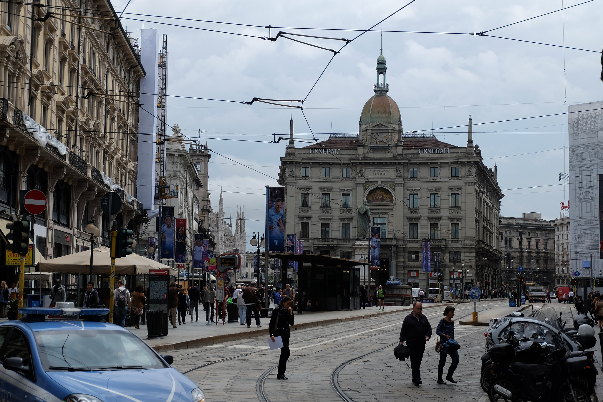 Eine belebte Stadtstraße mit einem parkenden Polizeiwagen, Fußgängern mit Taschen, fahrenden Fahrzeugen, Gebäuden mit Fenstern, Schildern, Laternen und Ampeln unter einem bewölkten Himmel.