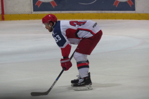 Hockey player in full gear skating on ice with a stick, facing a bannered wall in the background.