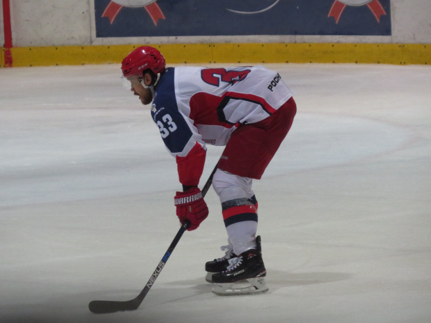 Hockey player in full gear skating on ice with a stick, facing a bannered wall in the background.
