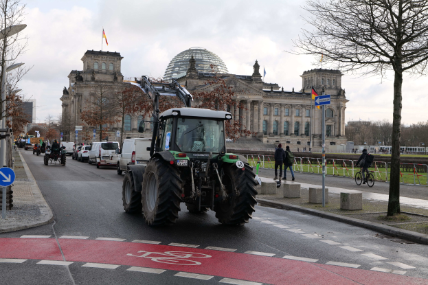 A tractor drives past the Reichstag building in Berlin, Germany, with pedestrians and cyclists on the sidewalk, trees lining the street, and flags adorning the building under a cloudy sky.