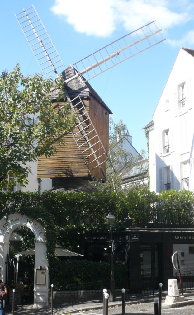 Eine Windm├╝hle, mehrere Geb├Ąude, B├Ąume, Metallstangen und ein Zaun in einer l├Ąndlichen Umgebung.