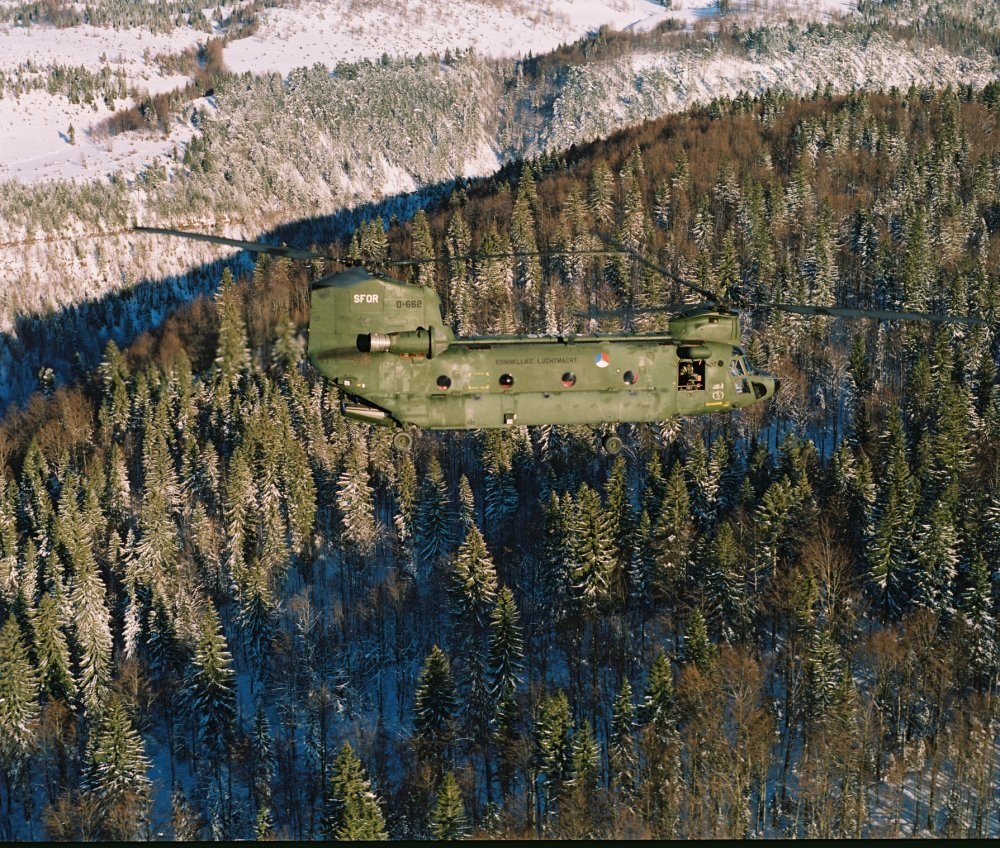 Militärhubschrauber fliegt über einen schneebedeckten Wald mit Bergen im Hintergrund, Text unten sichtbar.