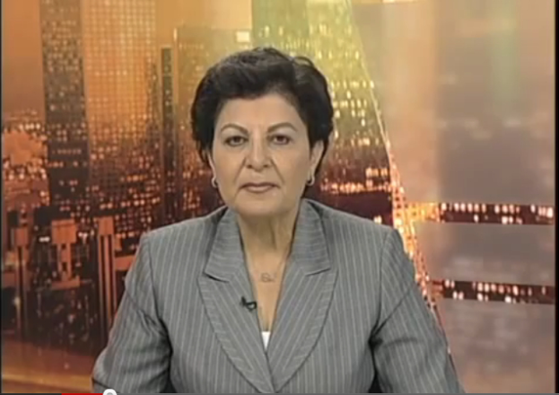 Eine Frau in einem Business-Outfit sitzt vor einem Fernseher mit Text am unteren Bildschirmrand, mit ein paar Gebäuden im Hintergrund.