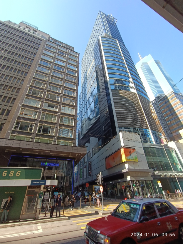 Roter Wagen auf einer Stadtstraße mit hohen Wolkenkratzern, Passanten auf dem Bürgersteig, Verkehrsampeln und sichtbare Straßenverkehrsschilder gegen einen klaren Himmel.