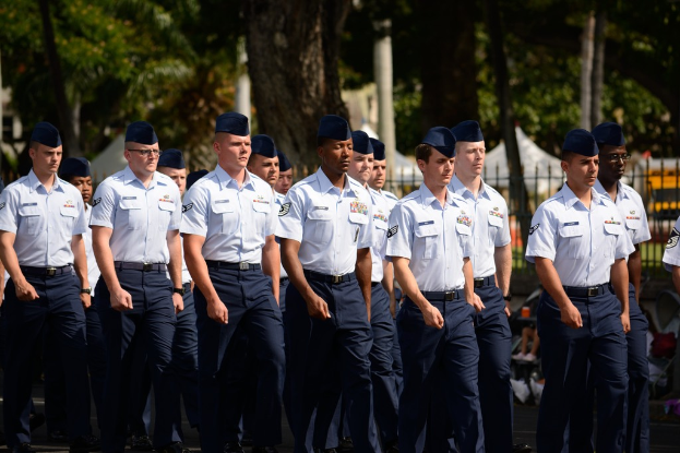 Männer in Uniform auf einer Straße mit einem Eisenzaun dahinter, Bäume und Pfosten im Hintergrund