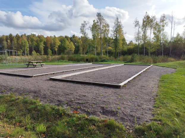 Eine grüne Wiese mit einem Picknicktisch, umgeben von Bäumen unter einem klaren blauen Himmel, mit Spielplatzgeräten wie Pfosten und einer Bank auf der linken Seite.