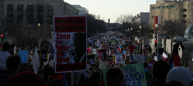 People protesting on a road holding posters with buildings, trees, light poles, and sky in the background.