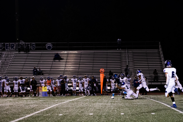 Ein Fußballspiel wird auf einem Rasenfeld bei Nacht gespielt, mit Spielern mit Helmen im Vordergrund und Zuschauern im Hintergrund.