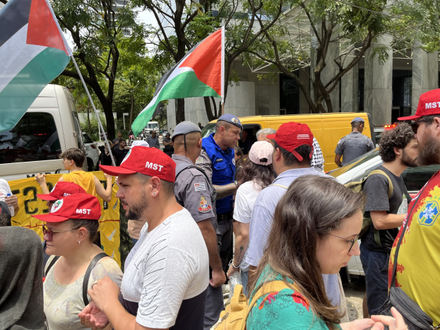 Eine Gruppe von Menschen mit palästinensischen Flaggen vor einem Gebäude mit Fahrzeugen und Bäumen im Hintergrund bei klarem blauem Himmel