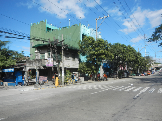 Eine Stadtstraße mit einem grünen Gebäude, fahrende Fahrzeuge, Fußgänger auf dem Gehweg und Strommasten mit Drähten bei einem bewölkten Himmel.