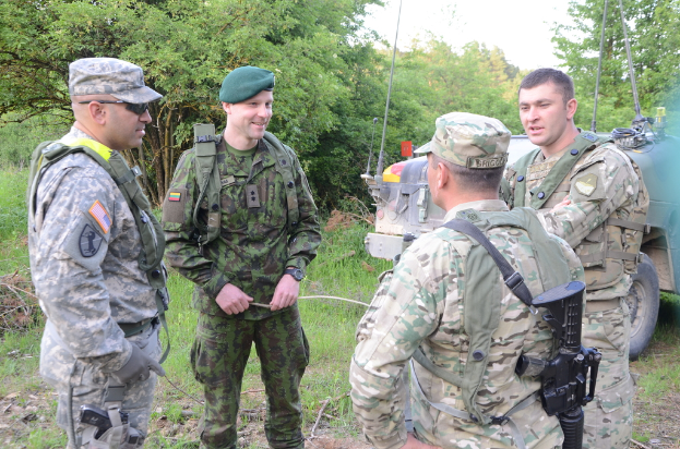 Vier Soldaten in Uniform, einschließlich Mützen und Waffen, stehen und sprechen, mit einem Fahrzeug rechts und Bäumen im Hintergrund.