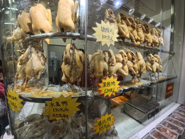 Vitrine mit ganzen gerösteten Hühnern auf einer Theke, eine Person links daneben, Deckenbeleuchtung im Hintergrund und Tabletts mit Lebensmitteln rechts.