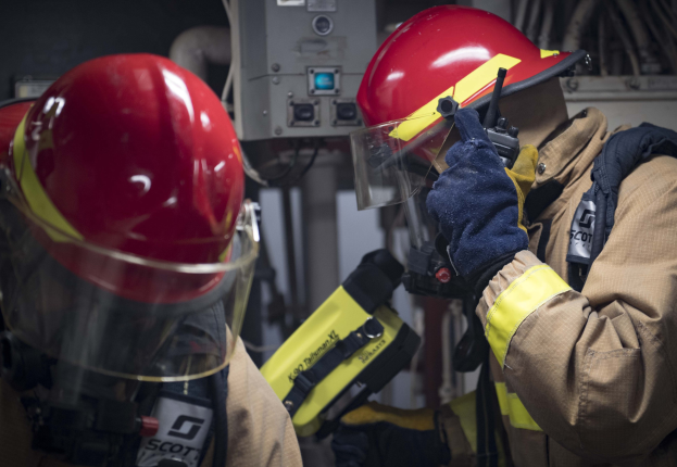 Zwei Feuerwehrleute in Schutzausrüstung arbeiten an einem Feuerhydranten während einer Ausbildung mit Maschinen und Kabeln im Hintergrund.