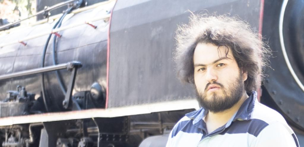 Ein Mann in einem T-Shirt steht neben einem Zug, mit sichtbaren Rädern und anderen Teilen des Zugs unten und einem Baum in der oberen linken Ecke.
