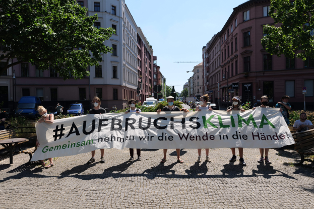 People in masks hold a banner reading "Aufbruchsklima" during a climate protest in Berlin, with surrounding urban elements and other participants visible.