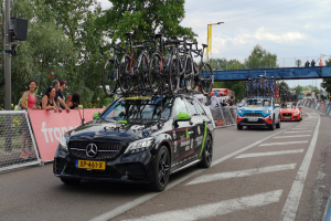 Ein Mercedes-Benz C63 AMG 4MATIC+ mit Fahrrädern auf dem Dach fährt eine Straße entlang, umgeben von Menschen, einem Zaun, einer Fahne, einem Straßenpfahl, einem Schild, einer Brücke, Bäumen, Drähten und einem bewölktem Himmel.