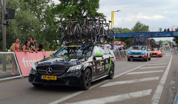 Ein Mercedes-Benz C63 AMG 4MATIC+ mit Fahrrädern auf dem Dach fährt eine Straße entlang, umgeben von Menschen, einem Zaun, einer Fahne, einem Straßenpfahl, einem Schild, einer Brücke, Bäumen, Drähten und einem bewölktem Himmel.