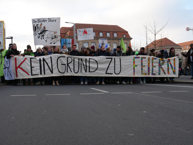 Protesters holding a banner reading "Kein Grund zu Feiern" against German austerity measures, with buildings, trees, and a clear sky in the background.