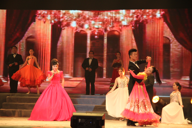 Eine Gruppe von Menschen auf einer Bühne, beleuchtet von Scheinwerfern, mit Stufen und einem Bildschirm im Hintergrund und einem Lautsprecher unten, was eine Tanzvorstellung vermuten lässt.